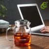 théière verre filtre avec filtre descendant et remontant remplie d'infusion, sur table en bois avec ordinateur en arrière-plan