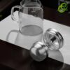 Théière verre transparente avec filtre en acier inoxydable à double mécanisme, posée sur surface texturée dans ambiance minimaliste