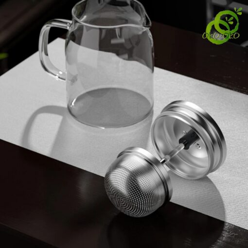 Théière verre transparente avec filtre en acier inoxydable à double mécanisme, posée sur surface texturée dans ambiance minimaliste
