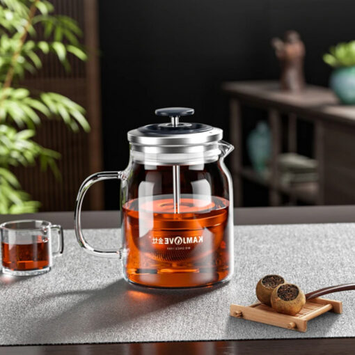 théière en verre transparent avec filtre rétractable intégré, remplie d'une infusion rouge foncé, posée sur une table avec une tasse et des théières de thé en boule