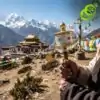 Molinillo de oración tibetano con mantras grabados | OkO-OKO™ Primer plano de una mano sosteniendo una rueda de oración tibetana dorada con mantras, frente a un fondo de picos nevados y un monasterio