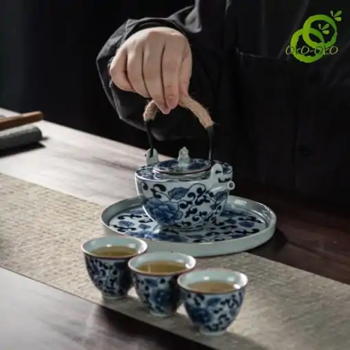 Blue and white porcelain tea set with a traditional lotus pattern | OkO-OKO™ A close-up of a teapot from a blue-and-white porcelain set with a lotus pattern on a wooden table, with three filled cups