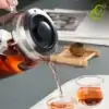 Glass Teapot with Retractable Filter | OkO-OKO™ Théière verre filtre en action, verse un thé chaud dans deux tasses, avec un couvercle noir et une poignée métallique, sur un plateau blanc