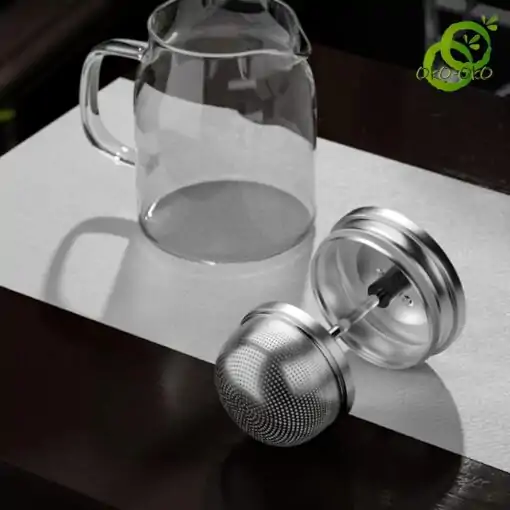 Glass Teapot with Retractable Filter | OkO-OKO™ Théière verre transparente avec filtre en acier inoxydable à double mécanisme, posée sur surface texturée dans ambiance minimaliste